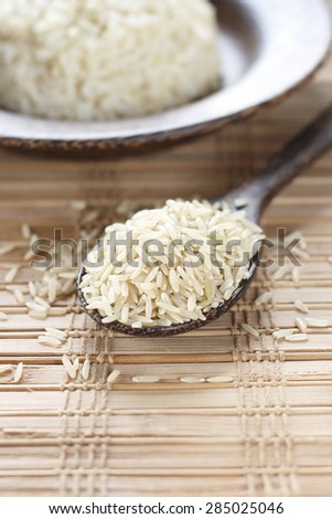 uncooked brown rice in wooden spoon and cooked brown rice in wooden plate in background.