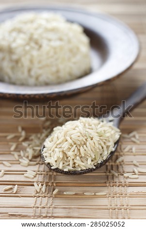 uncooked brown rice in wooden spoon and cooked brown rice in wooden plate in background.