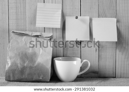 black and white picture style of paper bag of some food and coffee cup and note pad paper or note paper on wooden background. breakfast time with planing.