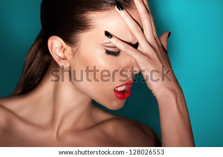 Fashion portrait of beautiful young woman with strong face expression. Touching her face with hand. Black nails manicure fashion makeup long black eyelashes red lipstick. Blue shaded background.