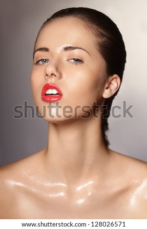 Fashion portrait of fresh young girl with shiny skin coated with oil. Natural make-up, beautiful blue eyes, tied brown hair, white teeth and red gloss lips. Gray shaded background.
