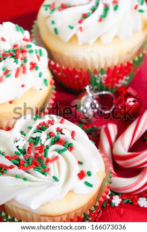 Holiday cupcakes with vanilla frosting and red and green sprinkles. Red holiday background with candy canes and bells. Closeup with shallow depth of field.