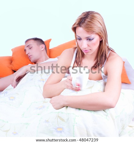 Young sad woman sitting on bed and drinking coffee, nearby slept  man. Focus on the woman.