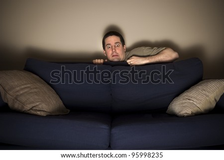 Frightened man hides behind couch while watching horror movie