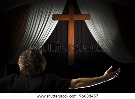A stock photo of a young man praying with open arms before a cross.