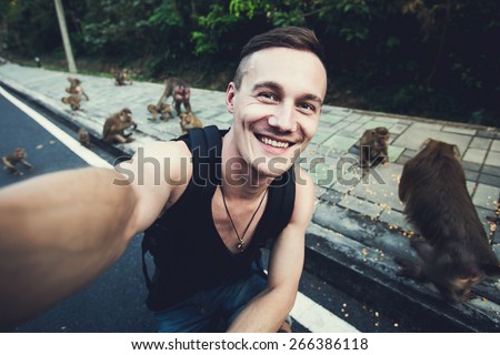 Handsome traveling man takes selfie photo with wild monkeys in tropical jungle forest in Phuket, Thailand, Asia