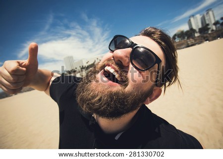 Handsome young bearded man making fun, laughing and posing with thumbs up on Santa Monica beach while travel in Los Angeles, California