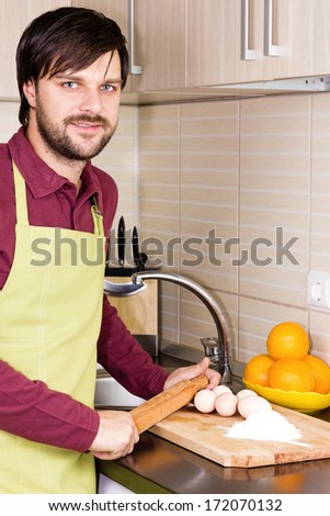 Smiling young man with apron holding a rolling pin ready to prepare a cake in his kitchen
