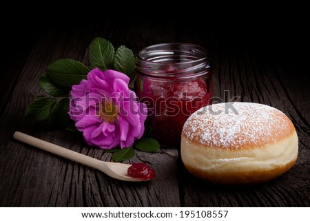 Fresh homemade donut covered by sugar powder frosting, wild rose flower and jar of red fruit jam. Sweets composition taken on rustic table.