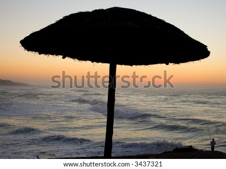 Silhouette of a grass umbrella with the sea in the background and the sun rising.
