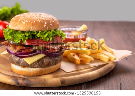 Hamburgers with fried bacon and sliced pickles  on a cutting board with french fries and tomato sauce
