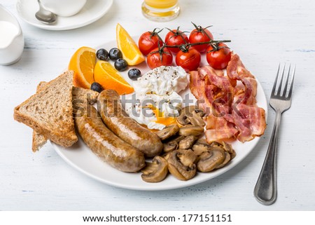 Healthy Full English Breakfast -  plate with poached eggs, sausages,  mushrooms, toasts and bacon, fruit, cup of fresh coffee and orange juice on white background