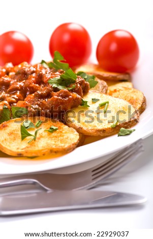 roasted country-styled potatoes with meat tomato sauce on a white plate, a fork and a knife and three cherry tomatoes at the back