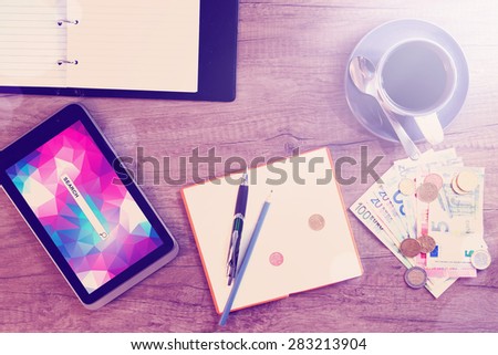 Digital tablet, sticky note paper, cup of coffee and money on old wooden desk. Simple workspace or coffee break with web surfing.