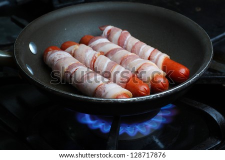 Wieners In A Blanket/Hot dogs wrapped in raw bacon frying in a pan on a stovetop with gas flames visible beneath
