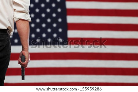 Man's in dark trousers & white shirt holding automatic pistol to one side with full view of out of focus US flag in background