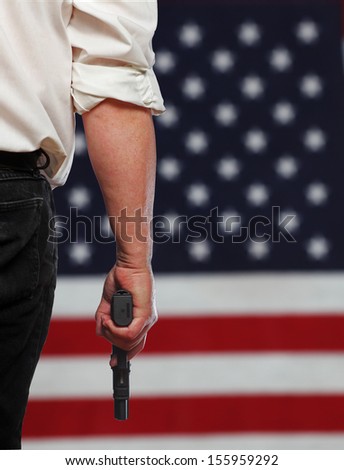 Close up of man in white shirt & dark trousers carrying handgun with out of focus cropped US flag in the background