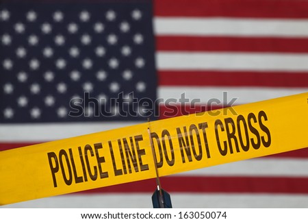 Crime scene barrier tape over out of focus US flag being cut with scissors