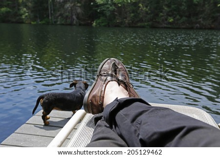 Man's feet & legs on lounge chair with small black dachshund & lake water in the background