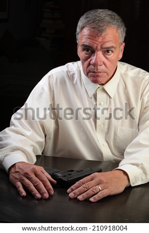Middle age man seated at table wearing white shirt with hands on table & automatic pistol set before him
