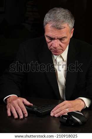 Middle age man seated at table looking down at table on which automatic pistol & folded tie are set