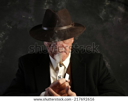 Man in suit jacket & fedora with brim over eyes lighting cigar with smoke pluming