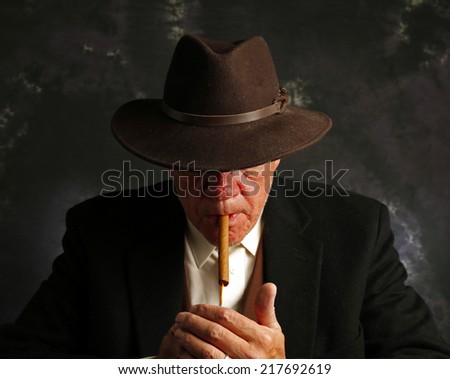 Man in suit jacket & fedora hat with brim over eyes lighting cigar