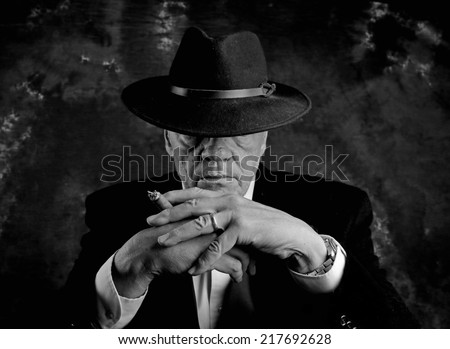 Black & white image of man in suit jacket & fedora with brim lowered over his eyes with lighted cigar