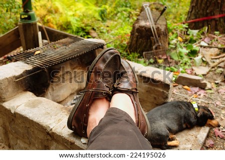 Man's feet in leather loafers resting on firepit with little dog napping in the background