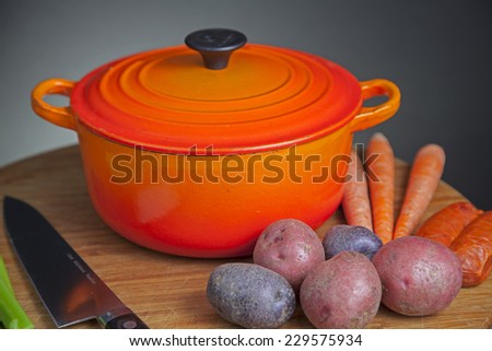 Orange enamel dutch oven on wood cutting board with potatoes, carrots, chef knife & celery arrayed