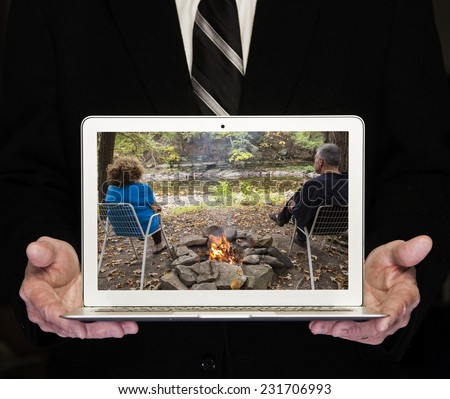Man in dark business suit & tie holding laptop computer with image of couple seated in the forest by a creek with a fire going.