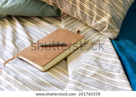 Unmade bed with pillows, sheets, custom journal & stainless steel pen set atop it.