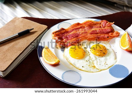 Breakfast in bed/Traditional breakfast of fried eggs & bacon on wood serving tray on unmade bed with journal & pen to one side