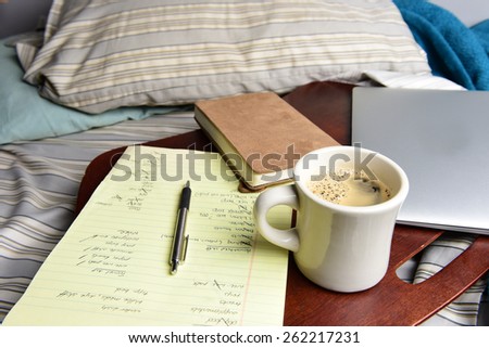 Home office/notebook computer, legal pad, pen on wood tray set on unmade bed with sheets & pillows & coffee mug