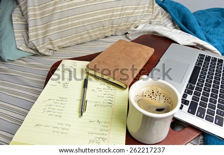 Home office/notebook computer, legal pad, pen on wood tray set on unmade bed with sheets & pillows & coffee mug,