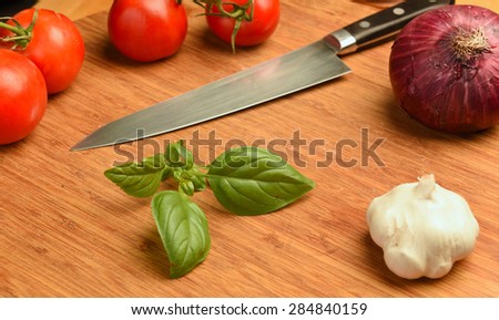 Fresh basil/cut basil sprig on cutting board surrounded by tomatoes, garlic & red onion with chef knife