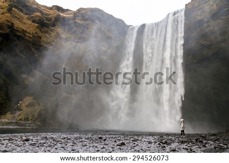 Beautiful female tourist dwarfed in front of the mighty Skogafoss waterfall in Iceland