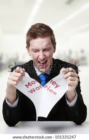 man in a suit sat at a desk ripping a  piece of paper up with the words money worries printed on it and looking happy doing so.