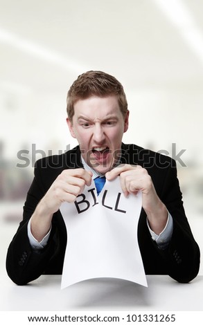man in a suit sat at a desk ripping a  piece of paper up with the word bill printed on it and looking happy doing so.