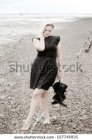 barefoot woman on a stoney beach wearing a dress and holding boots posing