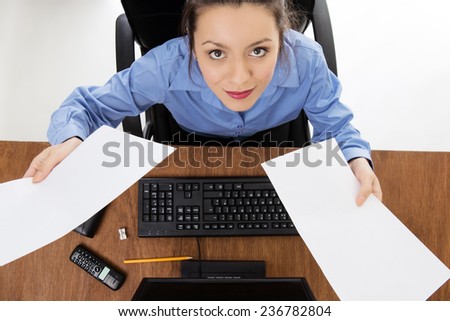 Woman sitting at her desk at work holding paper up in the air. Image is shot from a birds eye view looking down.