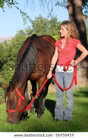 Young woman holding red lead rope while bay colored horse grazes on green grass.