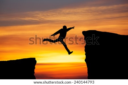 Man jumping across the gap from one rock to cling to the other. Man jumping over rocks with gap on sunset fiery background. Element of design.