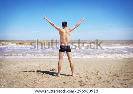 Back view of Young man spread his hands on beach. Man on beach hold hands arms up, rear view guy, standing back looking to sea blue sky horizon, vacation concept of freedom traveler