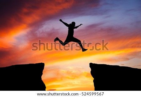 Man jumping across the gap from one rock to cling to the other. Man jumping over rocks with gap on sunset fiery background. Element of design.
