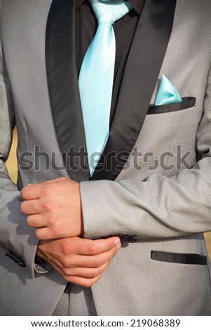 Caucasian man in a silver-grey formal suit with black trimmings. His hands are shown in front of his waist. He is wearing a turquoise tie and pocket handkerchief.