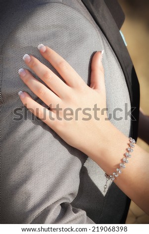 Young manicured hand of a woman on the arm of a man in a for lam suite. The woman is wearing a jewellery bracelet on her left hand