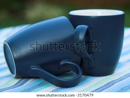 two blue mugs on a striped tablecloth. One mug is on it\'s side and the other is standing upright.  The mugs are blue. The tablecloth blue, green and white stripes.