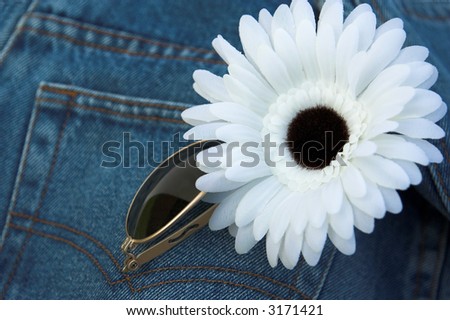 Blue denims with sunglasses and a flower on the back pocket. Focus is on the flower, Denims are blue. Sunglasses have a gold frame. Flower is a daisy and is made of white material.