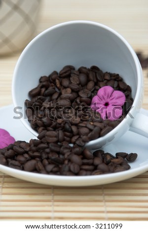 White cup lying on a saucer with coffee beans spilling out. There is a pink flower in the cup.  The beans are pouring put towards the viewer